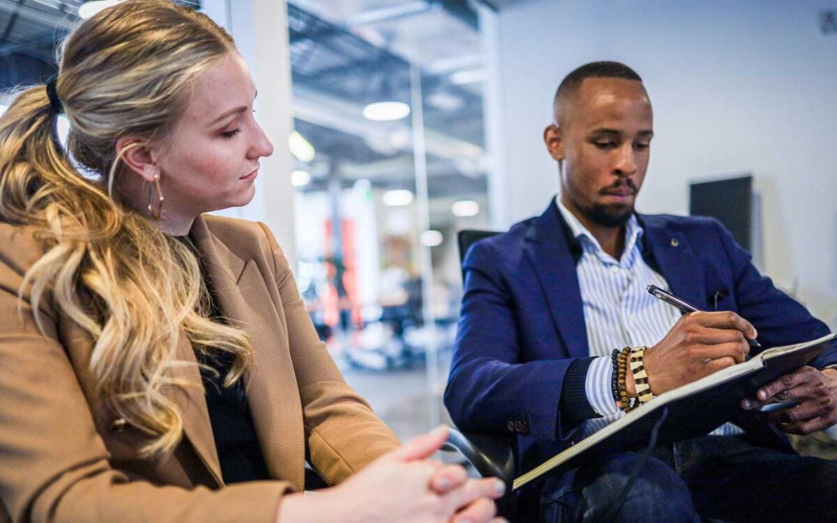 Man sitting next to woman, writing in a notebook in a business setting