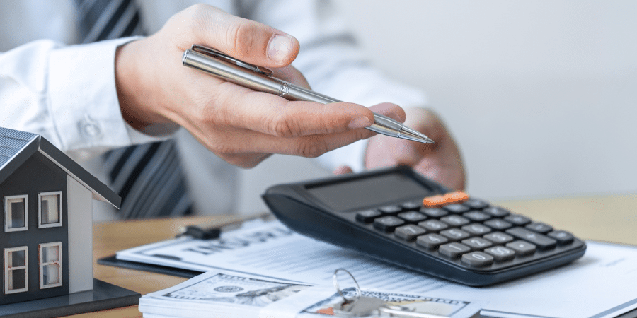 A man holding a pen and showing numbers on a calculator, sitting on a stack of papers.