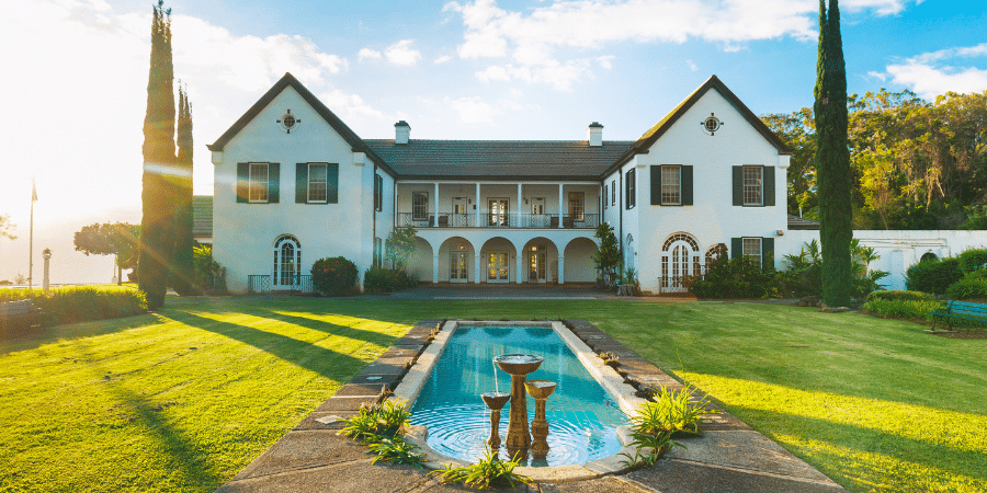 Photo of exceptionally large house framed by large skinny pine trees with a reflecting pool in front in the center of a plush green lawn.