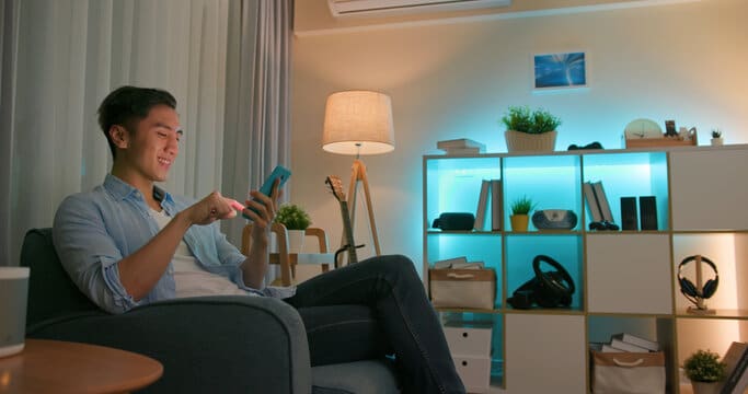 Smiling man on his phone, his shelving is lit up in blue, with soft lights in his living room.