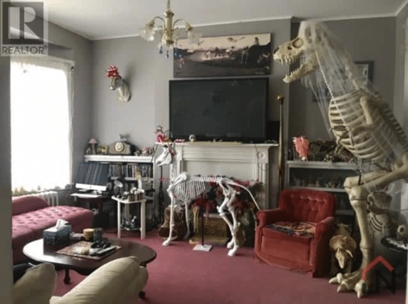 A sitting room with a red tufted bench and chair with a skeleton of a T. Rex wearing a wedding veil standing on one side of the room. There is also a skeleton of a large breed dog in front of the fireplace.