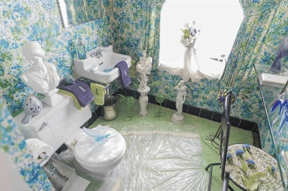 Bathroom with blue and green floral wallpaper and curtains to match. There are several white sculptures standing, including one bust on the back of the toilet. The toilet lid is adorned with a large butterfly and there's a large plastic sheet on the floor wrapped around the base of the toilet.