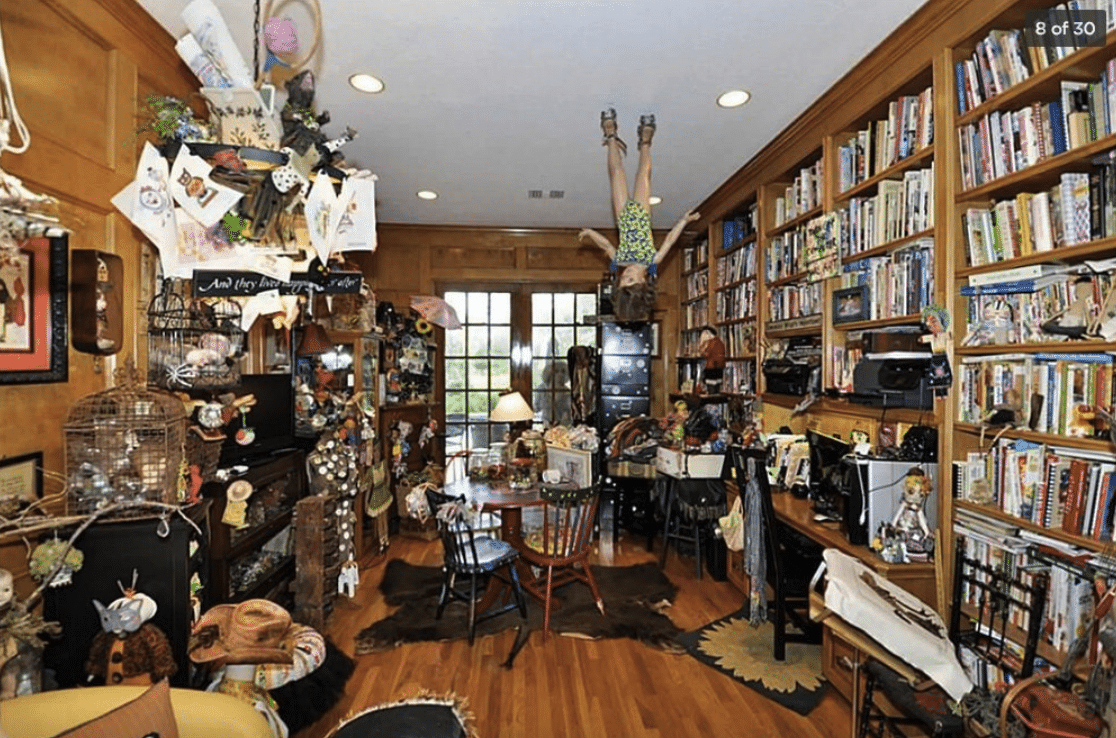 Library with floor-to-ceiling bookshelves on one wall, a table with three chairs, tons of additional clutter on the opposite side of the room, and a mannequin child hanging upside down from the ceiling.