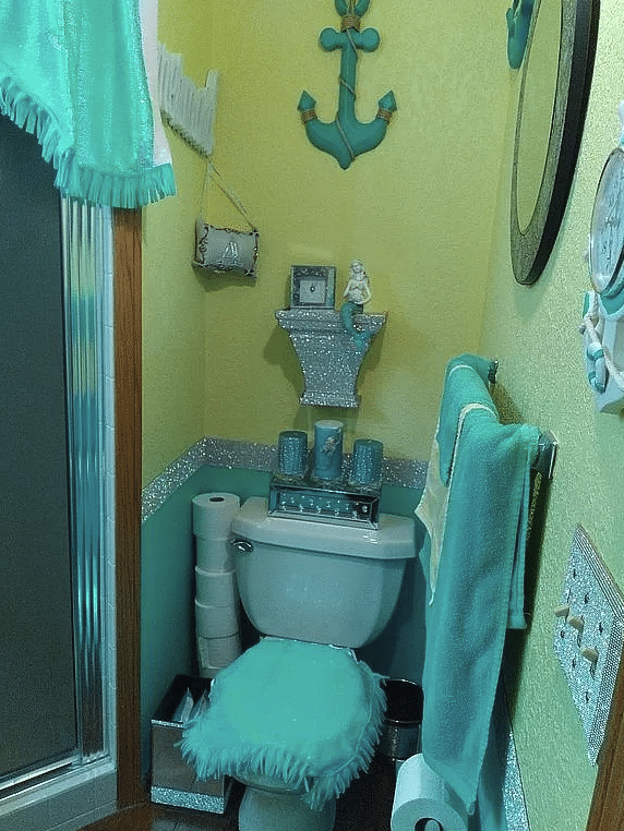 A small bathroom with an aqua-colored toilet, aqua, and lime-colored walls with a strip of glitter between the two colors. The light switch plate is also glitter and the toilet lid is covered with an aqua-colored fuzzy cover.