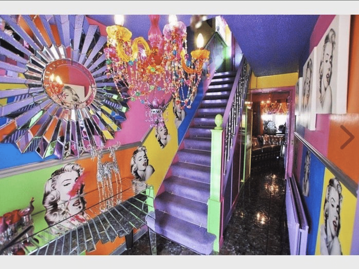 A hallway with a staircase covered in purple carpeting. The walls in the hallway are multi-colored in colored blocks with images of Marilyn Monroe all over them and a multi-colored chandelier hanging from the ceiling.