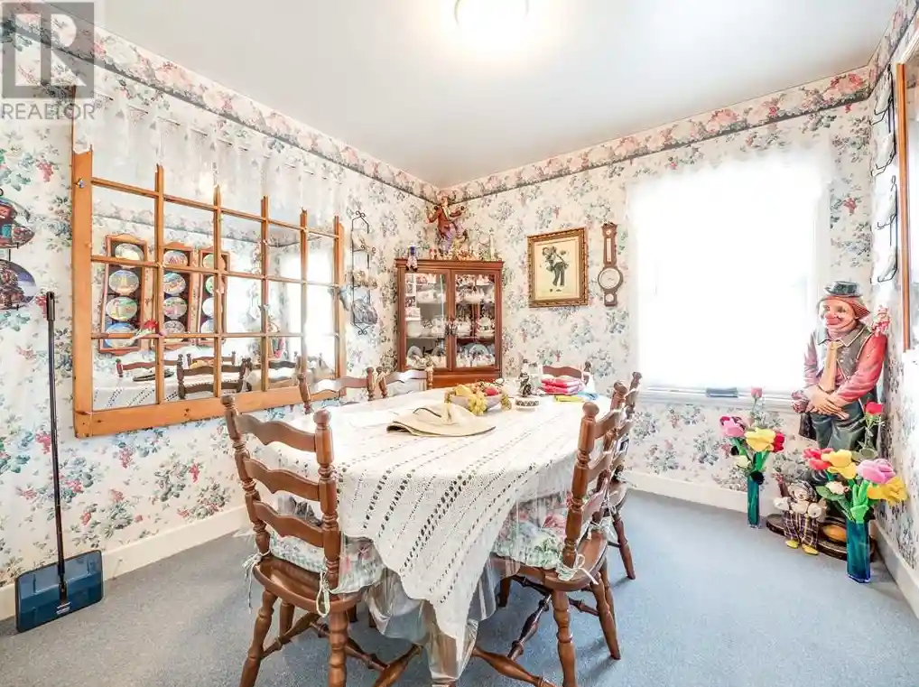 Small dining room with floral wallpaper, a large mirror, a wooden table with ladder-back chairs, and a four-foot-tall clown standing in the corner surrounded by vases holding multi-colored flowers.