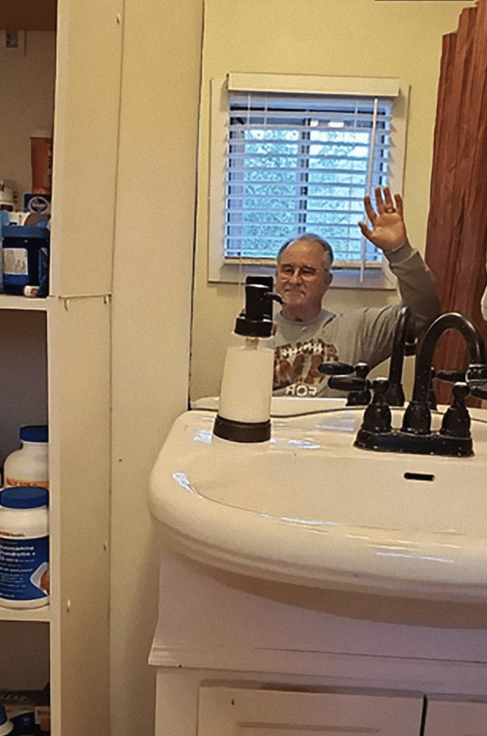 A man sitting on a toilet waving into a mirror over the bathroom sink.