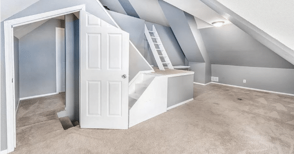 A room whose door is cut on a slant at the top, in front of a set of stairs that lead to a small landing and another set of stairs that appear to lead to a crawl space. There is an additional ladder that also leads to the crawl space. The room is full of 45-degree angles on the walls and ceiling.