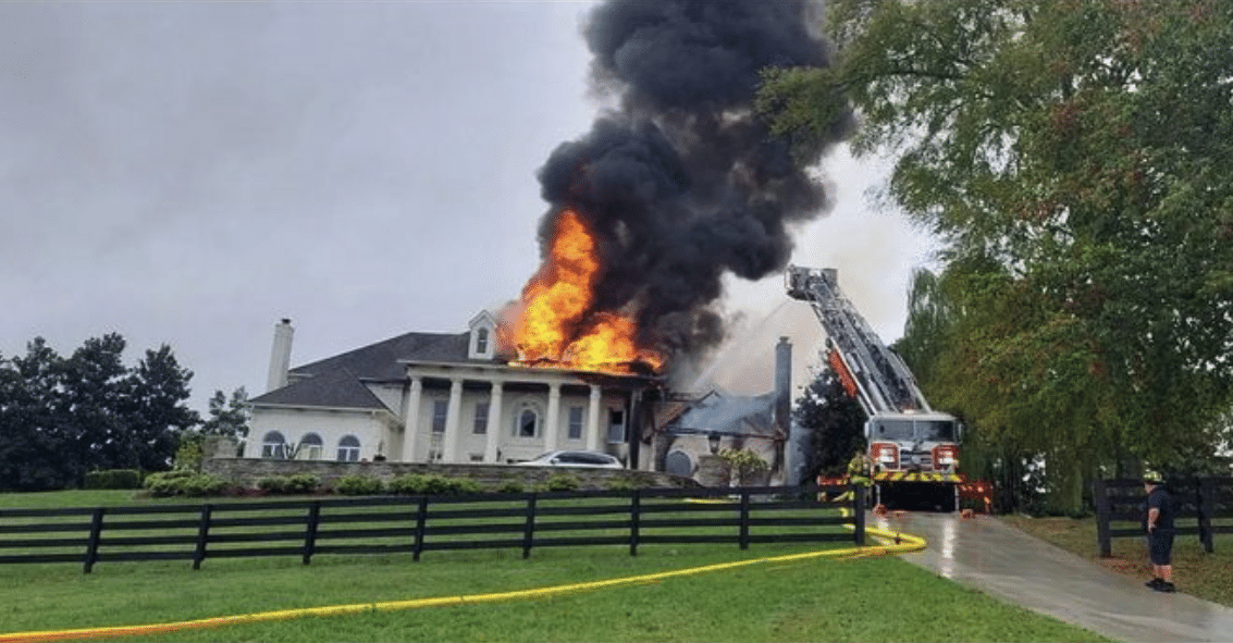 A large mansion set back from the road with flames pouring from the top with plumes of black smoke and a fire engine parked in the drive with firemen trying to extinguish the fire with their water hoses.