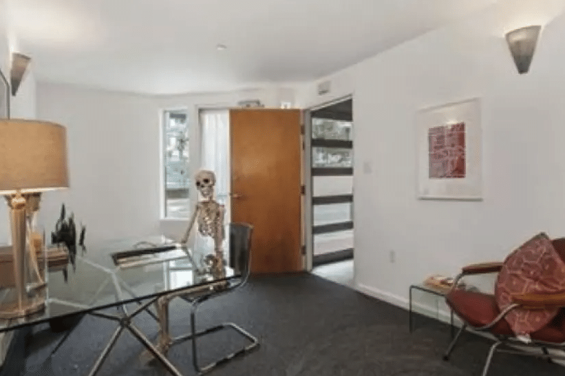 A room with a chair and a glass-top table with a skeleton sitting at the table.