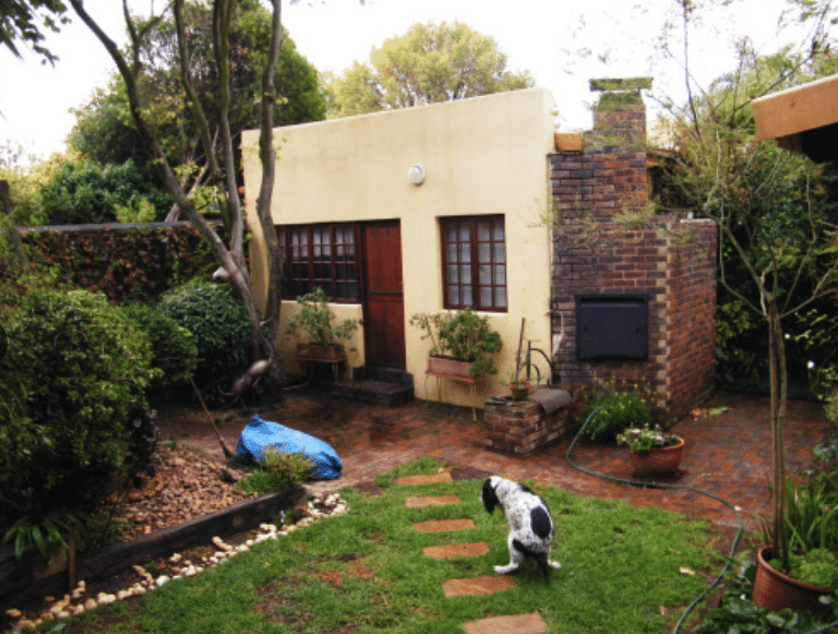 Exterior of a cottage home and a well-manicured garden with a dog pooping on the green grass next to the stone walkway.