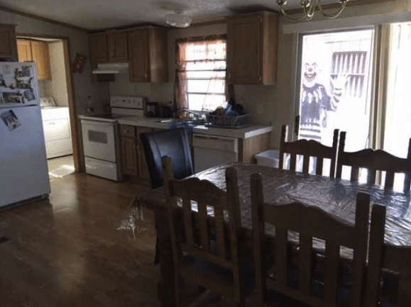 Dark photo of a kitchen and portion of a dining room with a large man dressed as a creepy clown standing just outside the glass patio door waving.