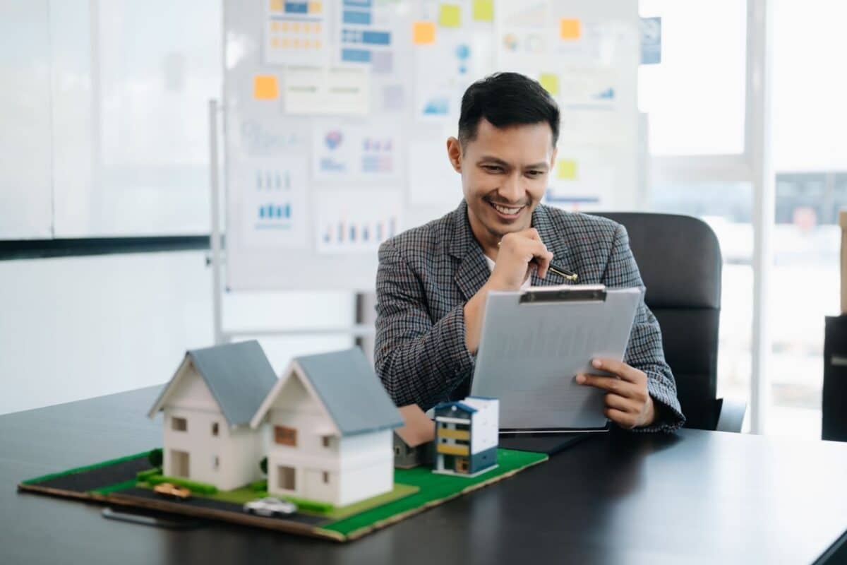 Young real estate agent worker working with laptop and tablet at table and small house beside it.