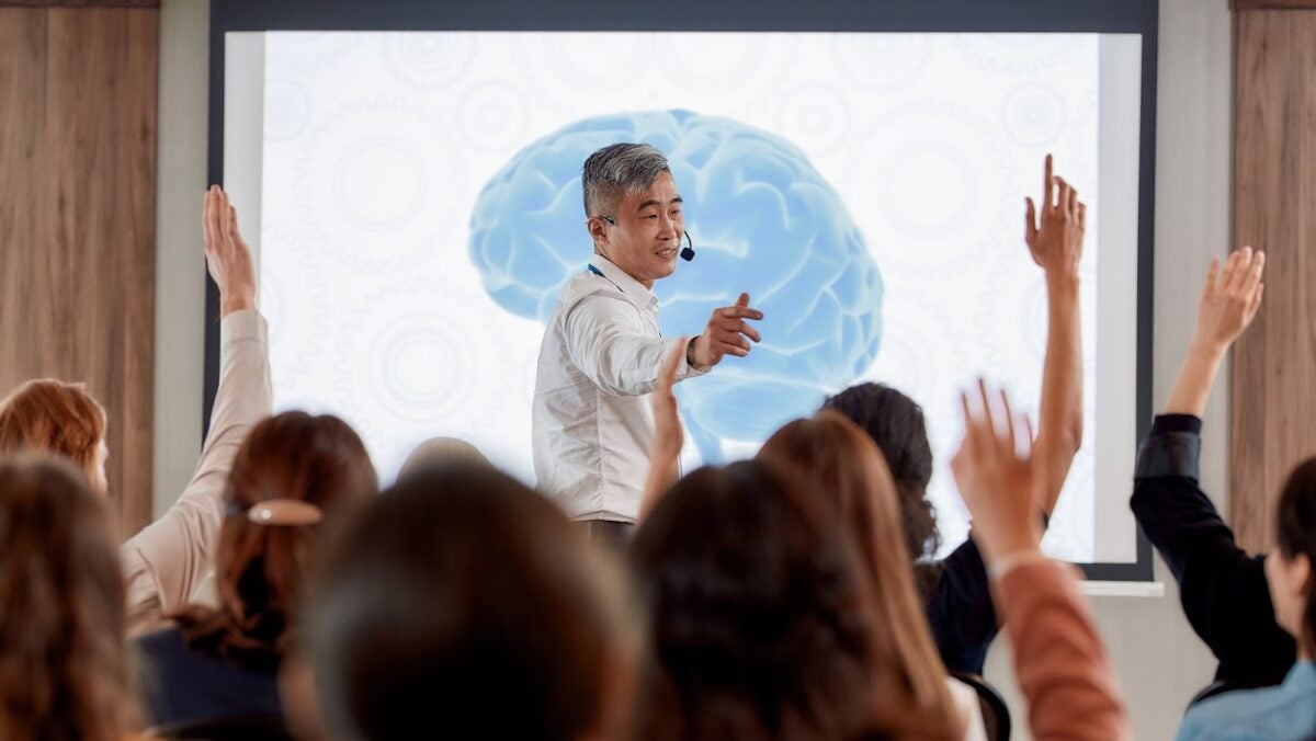 A mentor wearing a headset and standing in front of a room full of people with raised hands.