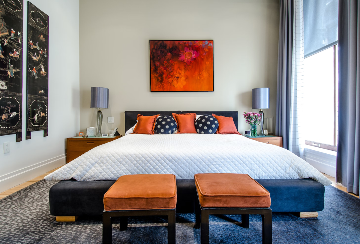 A small carpeted bedroom with floor-to-ceiling curtains and a white bed with orange and dark blue pillows