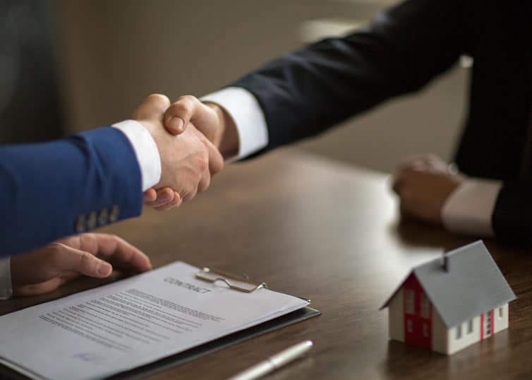 Two people sharing a handshake over a table with a contract on it.