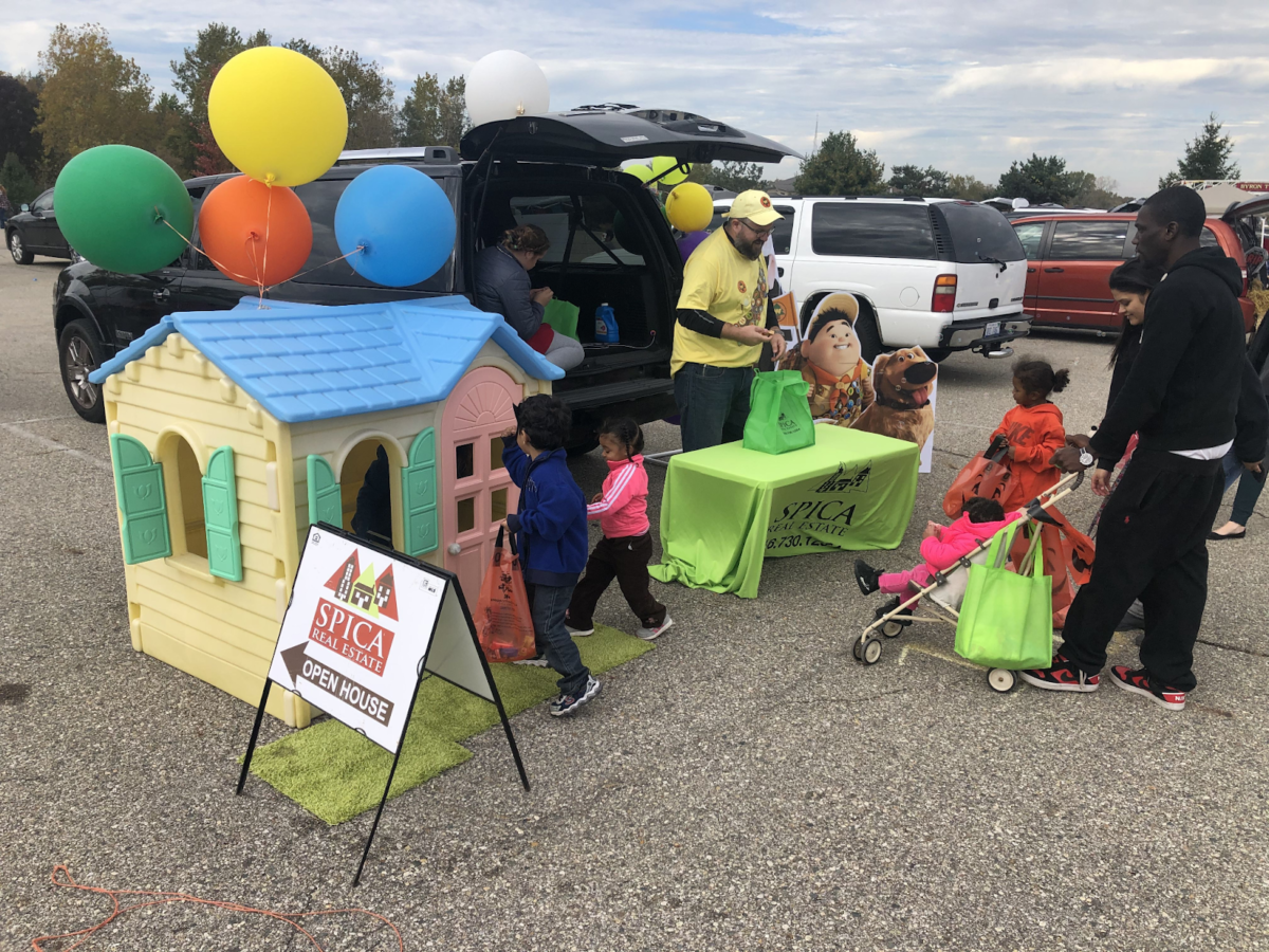 A real estate agent at a trunk or treat event passing out candy with a play house and an open house sign in front of it.