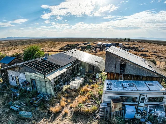 Two structures sitting next to each other with a camper in front of one. The structures are falling apart, and the roof is missing in multiple places. The structures are situated in a field that is covered in debris.