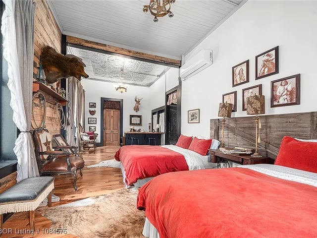 Inside a hotel room with two red beds. The room has an animal-skin rug and a taxidermied bear head hanging above the fireplace. Between the two beds hanging on the walls are vintage photographs.